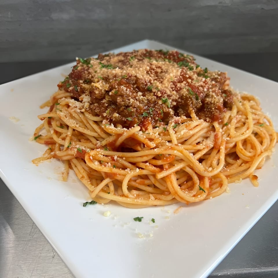 Spaghetti with meat sauce, parmesan cheese, and parsley on a white square plate.